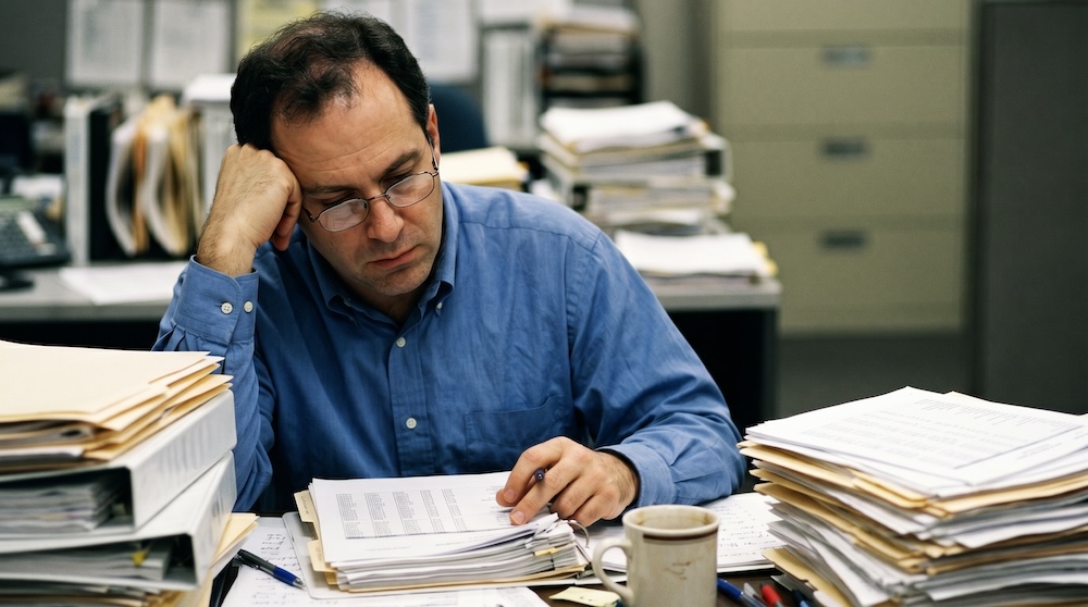 Overworked accounting employee processing paper invoices manually at a cluttered office desk. Stressed finance professional reviewing stacks of documents, highlighting the hidden costs, delays, and inefficiencies of manual AP/AR workflows and outdated invoice processing systems in a business environment.